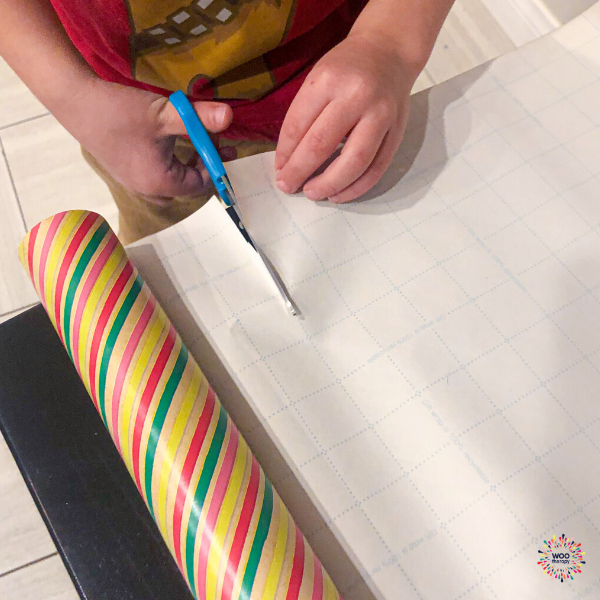 Child using appropriate scissors and guide marks to cut paper in preparation for gift wrapping.